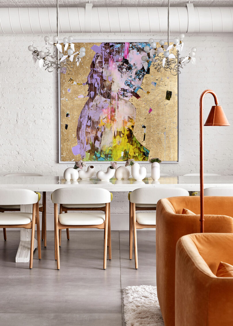 A modern dining area with a white table, wooden chairs, and an abstract portrait on the wall. Orange armchairs and a unique chandelier add contrast to the minimalist space.