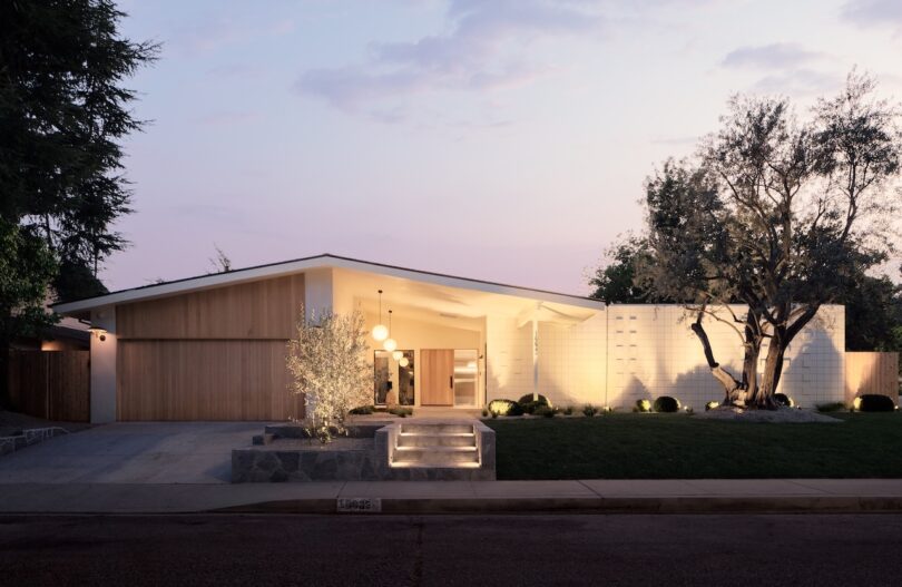 A modern, single-story house with a flat roof, wood and white exterior, front yard landscaping, and outdoor lighting at dusk.