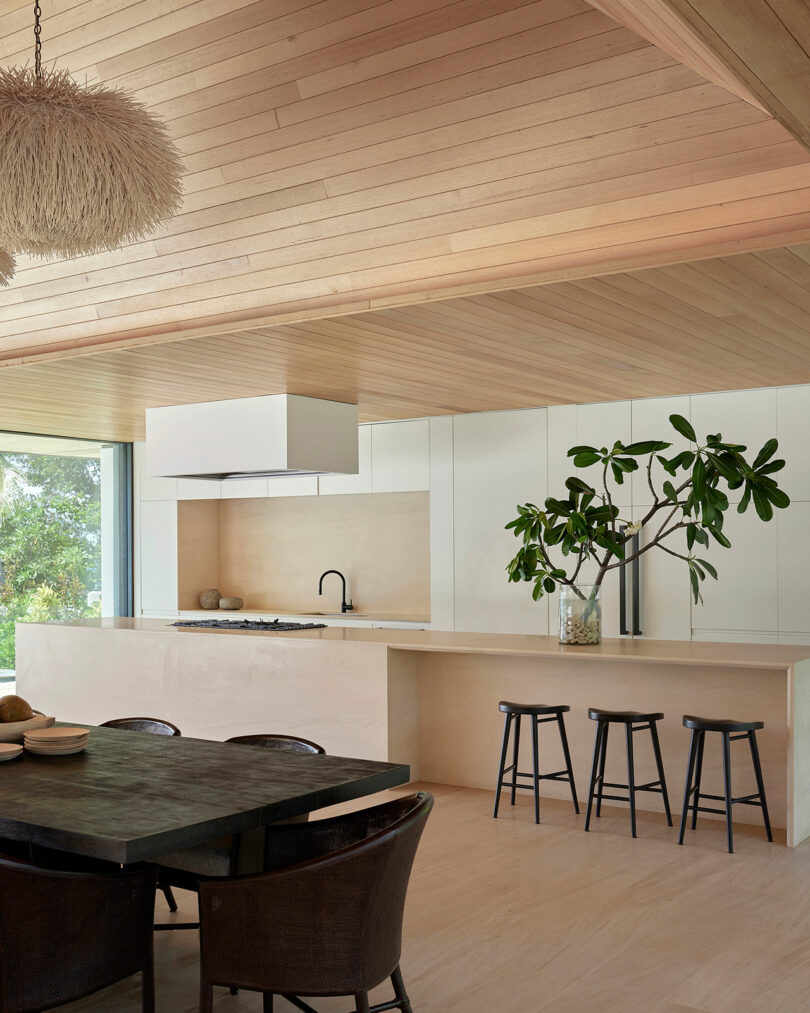 Modern kitchen with a light wood finish, island counter with three black stools, a plant in a glass vase, and a dark dining table with chairs. Ceiling features a wooden design and a chandelier.