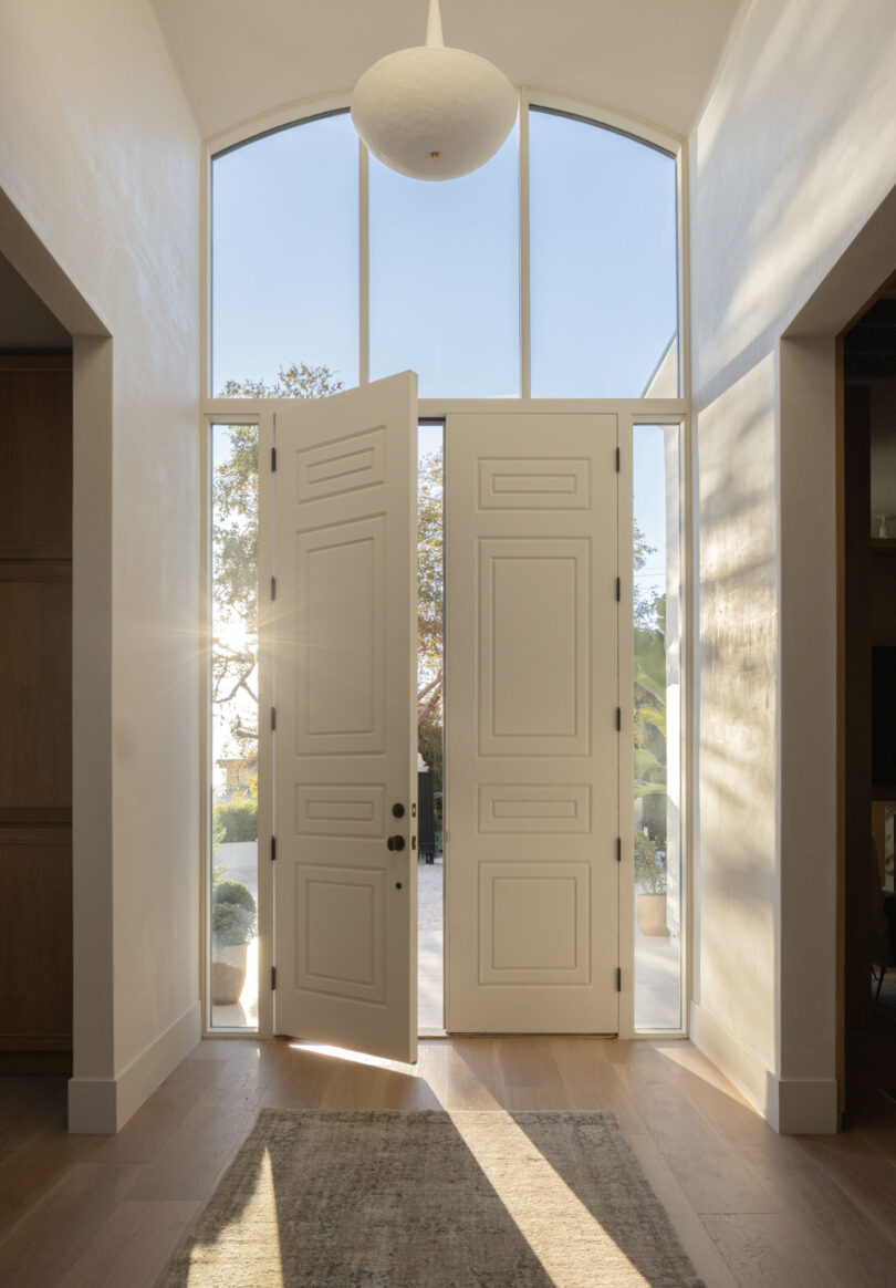 A sunlit entryway featuring double doors, tall windows, and a modern light fixture. The doors are open, revealing an outdoor space with trees and plants.