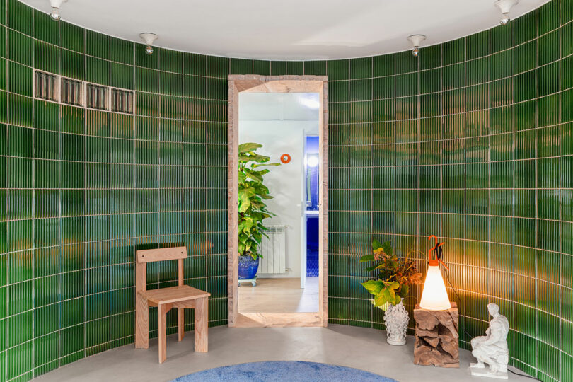 Room with green tiled walls, a wooden chair, plants, a standing lamp, and a statue. A central doorway leads to a space with more plants and a blue wall.